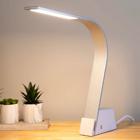 A Brooklyn AC LED Task Lamp on a table next to succulents. The lights' 3000K glow is illuminating the desktop surface, and the USB-A port is exposed.