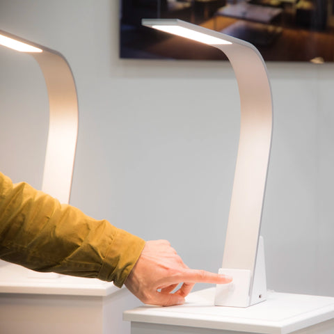 Hand using the touch-activated front panel of the Brooklyn USB LED Task Lamp in Brushed Aluminum. The USB-A port is visible.