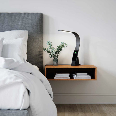 Brooklyn Aura LED Task Lamp on a floating wooden night stand next to a bed. There is a phone wirelessly charging on the Brooklyn Aura LED Task Lamp's device shelf.