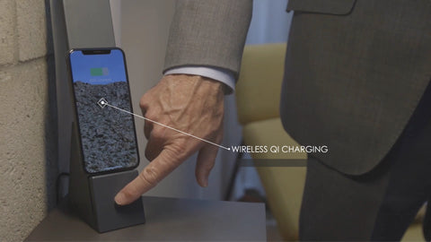 A man in a business suit using his finger to touch-activate the Brooklyn AURA LED Task Lamp in Black Slate. The lamp has an iPhone being charged wirelessly. There is text on top of the image that reads "Wireless Qi Charging"