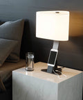 Brooklyn USB LED Desk Light in Black Slate finish on a contemporary bedside table next to a glass of water. The light has an iPhone being charged via USB-A port resting on its device shelf. The USB cord is hidden behind the device shelf. 