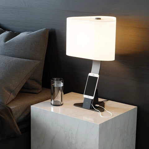 Brooklyn USB LED Desk Light in Black Slate finish on a contemporary bedside table next to a glass of water. The light has an iPhone being charged via USB-A port resting on its device shelf. The USB cord is hidden behind the device shelf. 