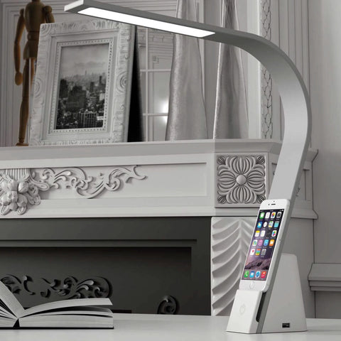 Close up of the Brooklyn Aura LED task Lamp on a desktop next to a book. There is a phone charging on the device shelf.