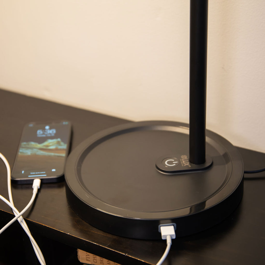 The base of a LUX Highline LED Desk Lamp charging a phone with one of its two USB charging ports built into the base.